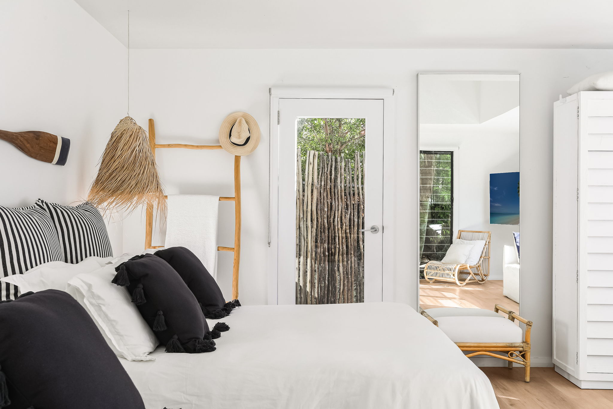 Bedroom with black tassel pillows, black-and-white stripe cushions, rattan pendant, straw hat on towel ladder and bamboo door