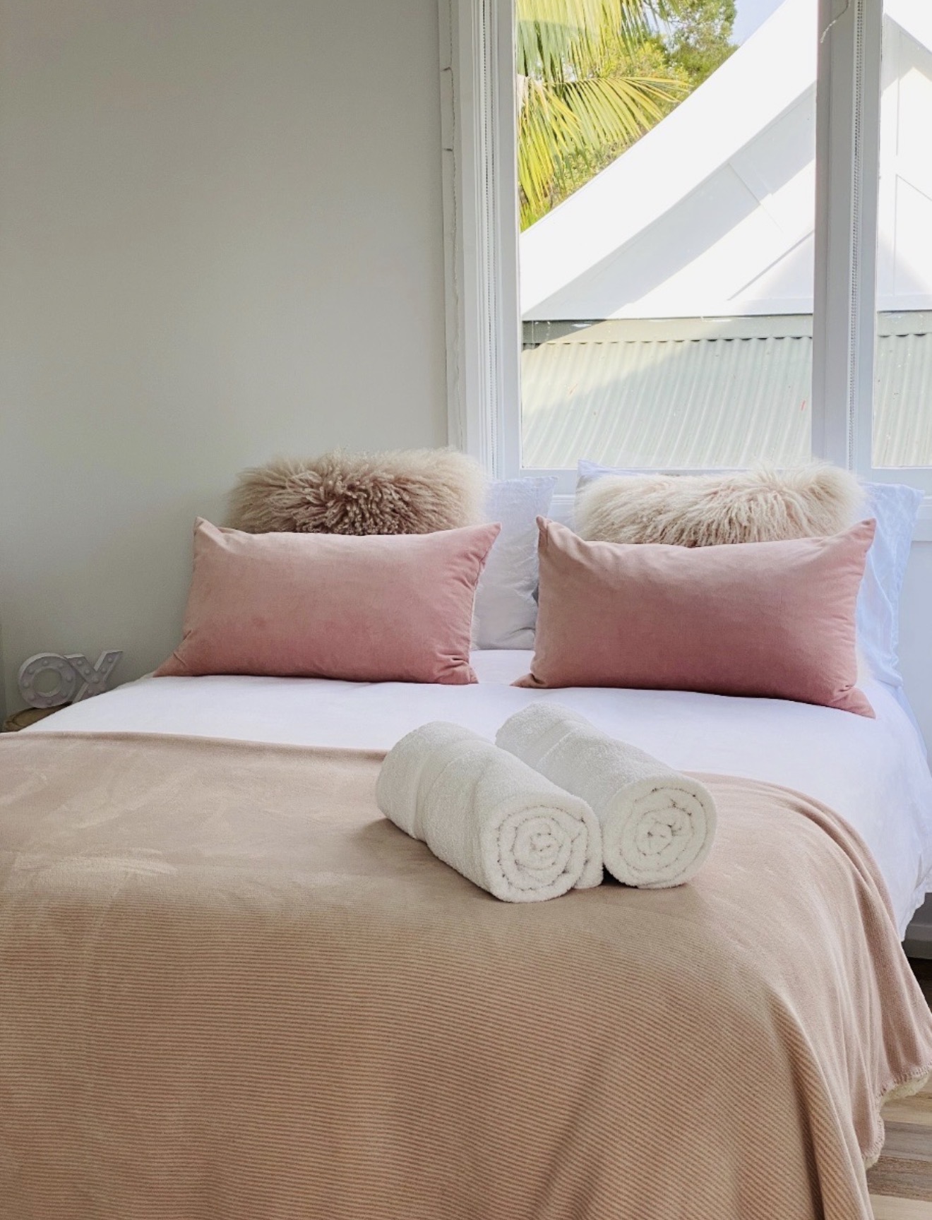 Bed with pink velvet pillows, mongolian fur cushions and rolled towels by a bright window