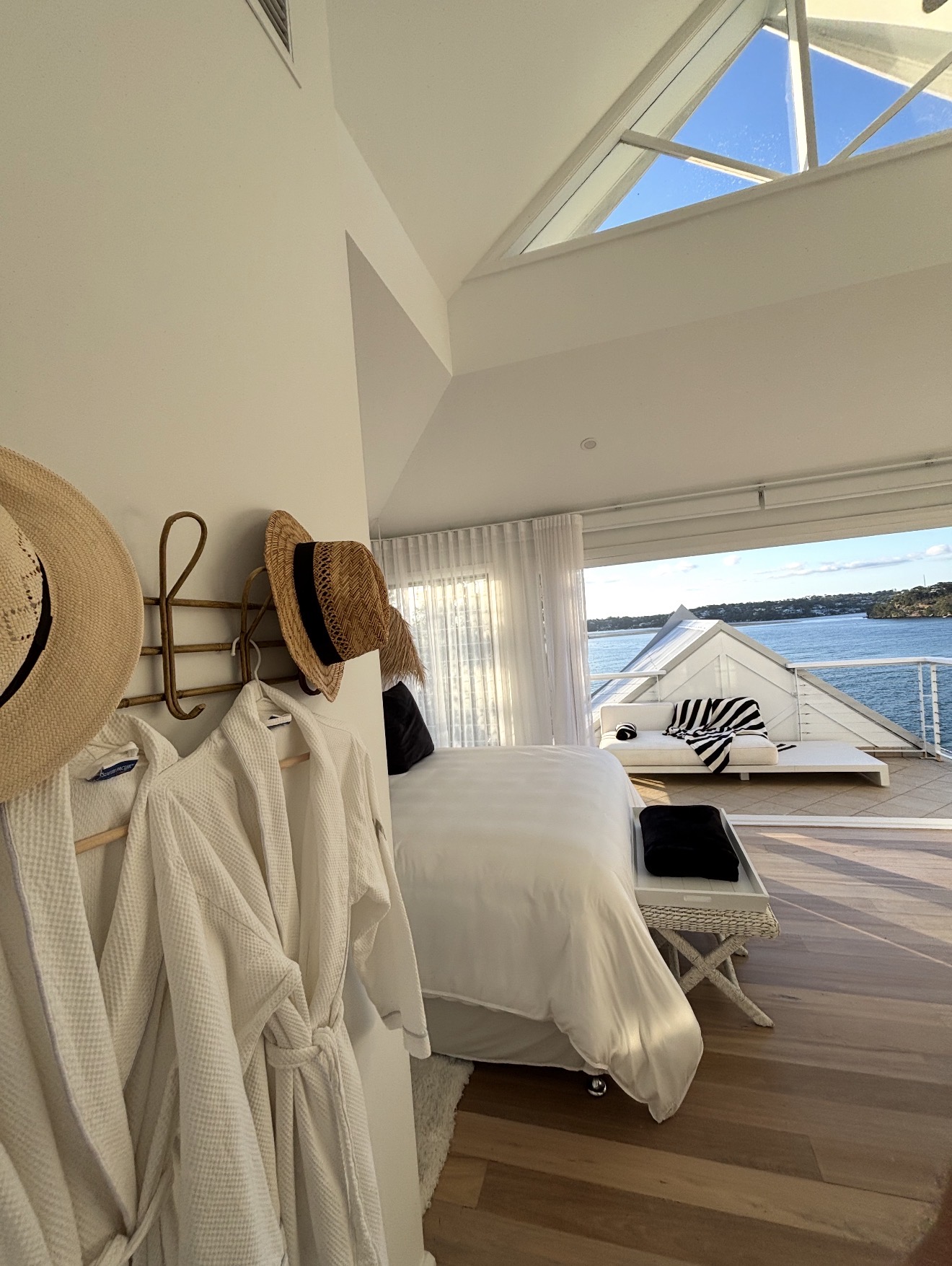 Bedroom with white robes and straw hats on wall hooks opening to a deck with bay views