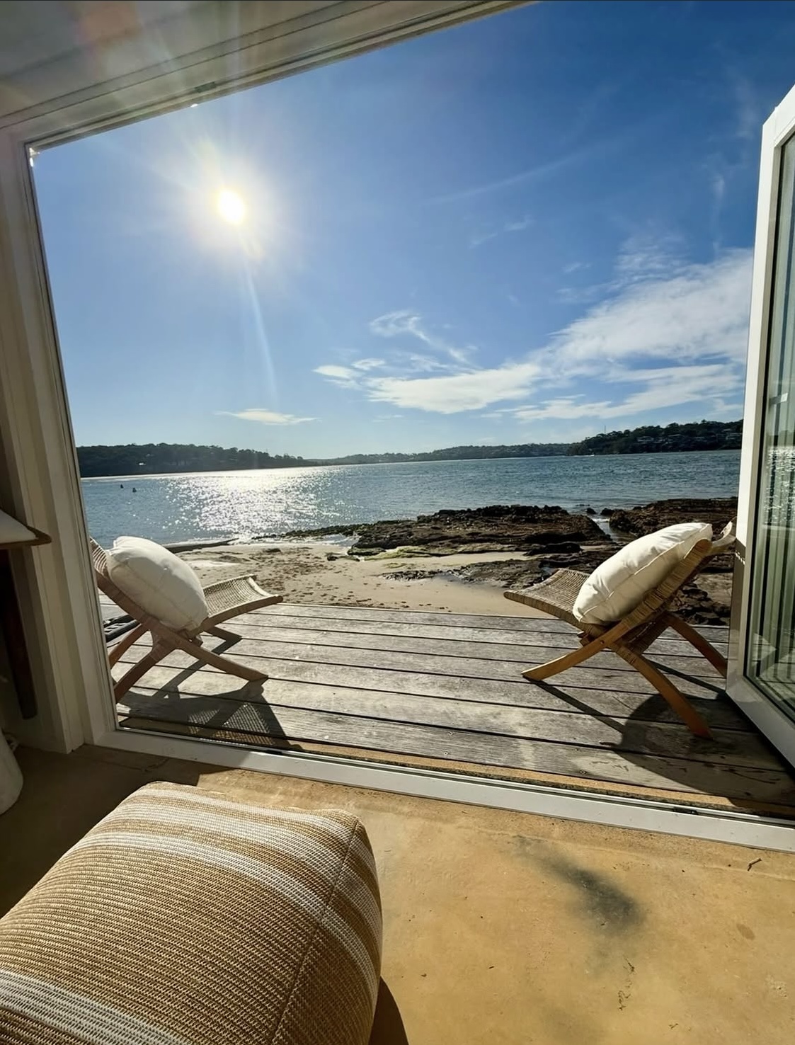 Two rattan sling chairs on a timber deck framed by open bifold doors with beach beyond