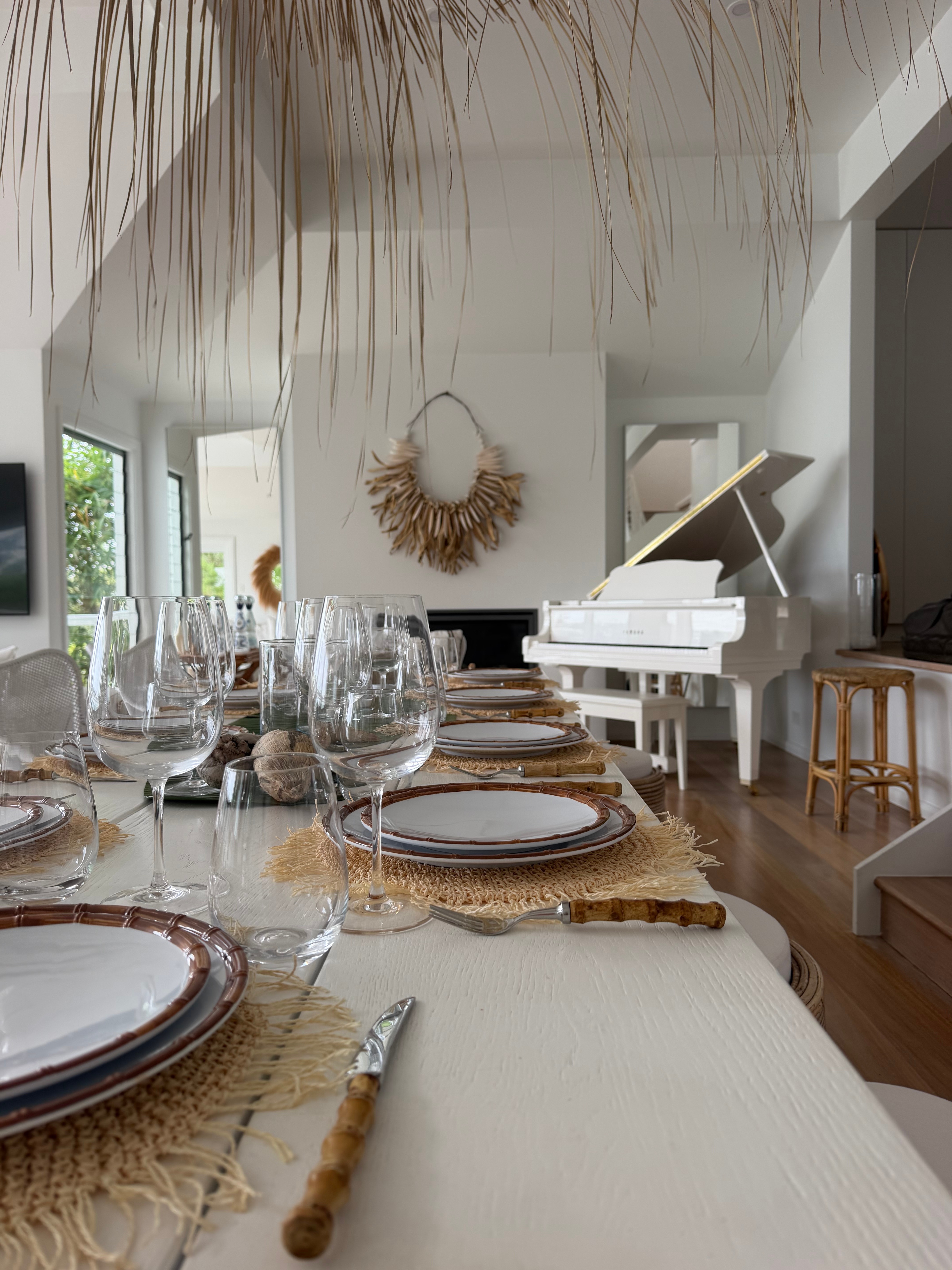 Long dining table set with wine glasses under dried palm fronds with white grand piano beyond