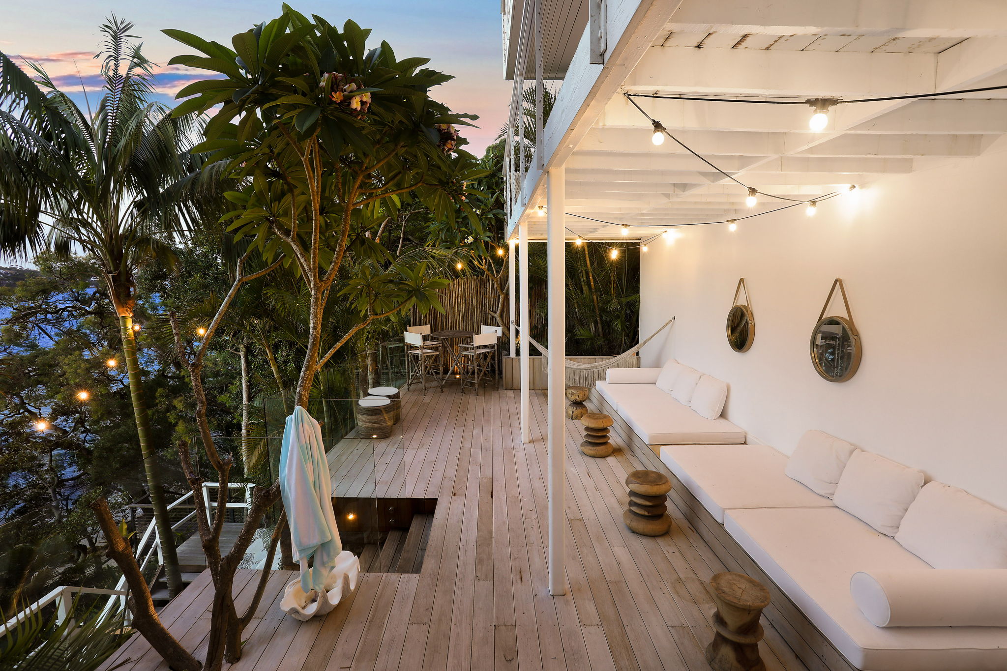 Dusk deck with festoon string lights, white built-in sofa bench, timber stools and frangipani tree