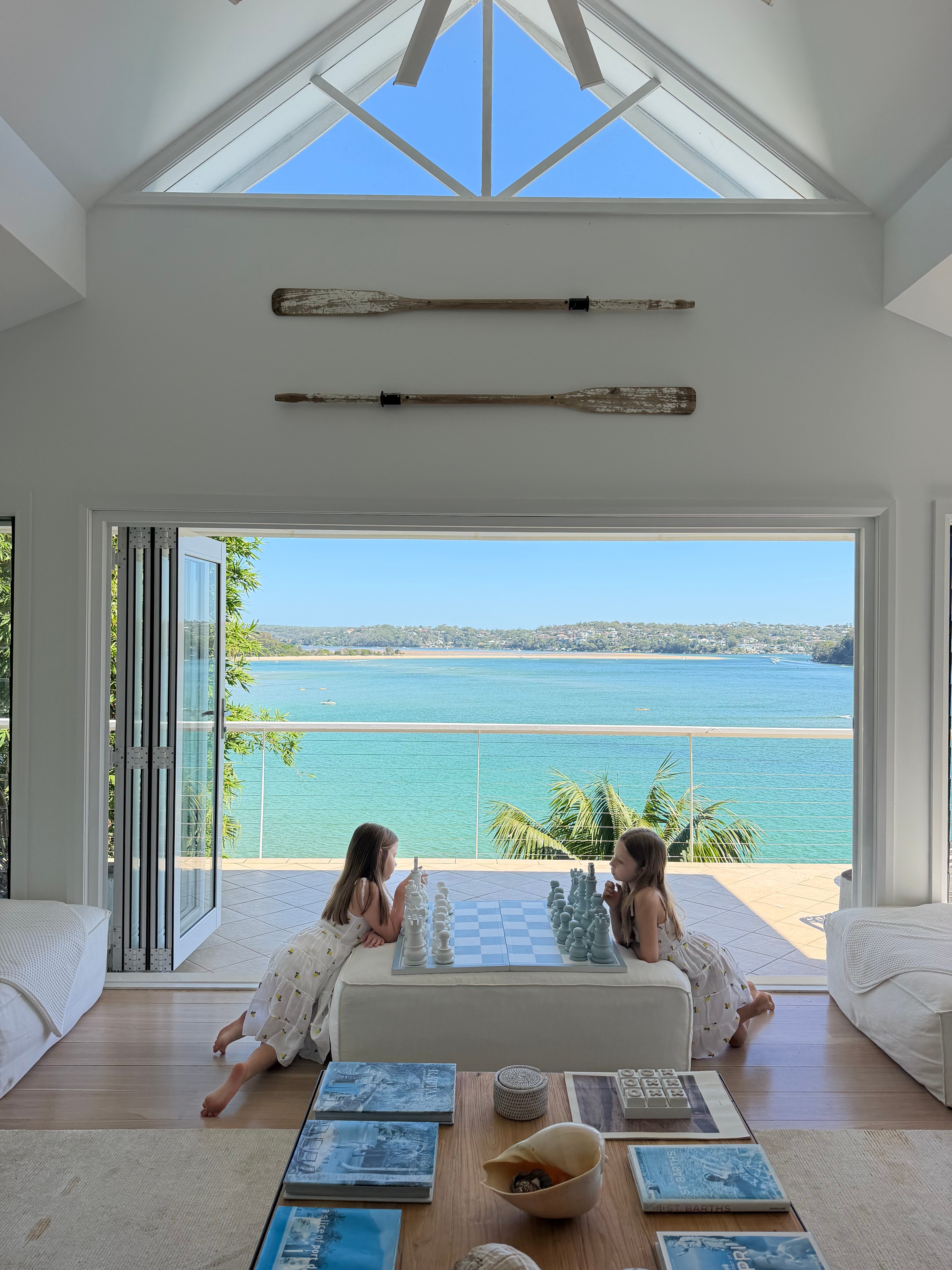 Two girls playing chess on white ottoman under cathedral ceiling with turquoise bay beyond