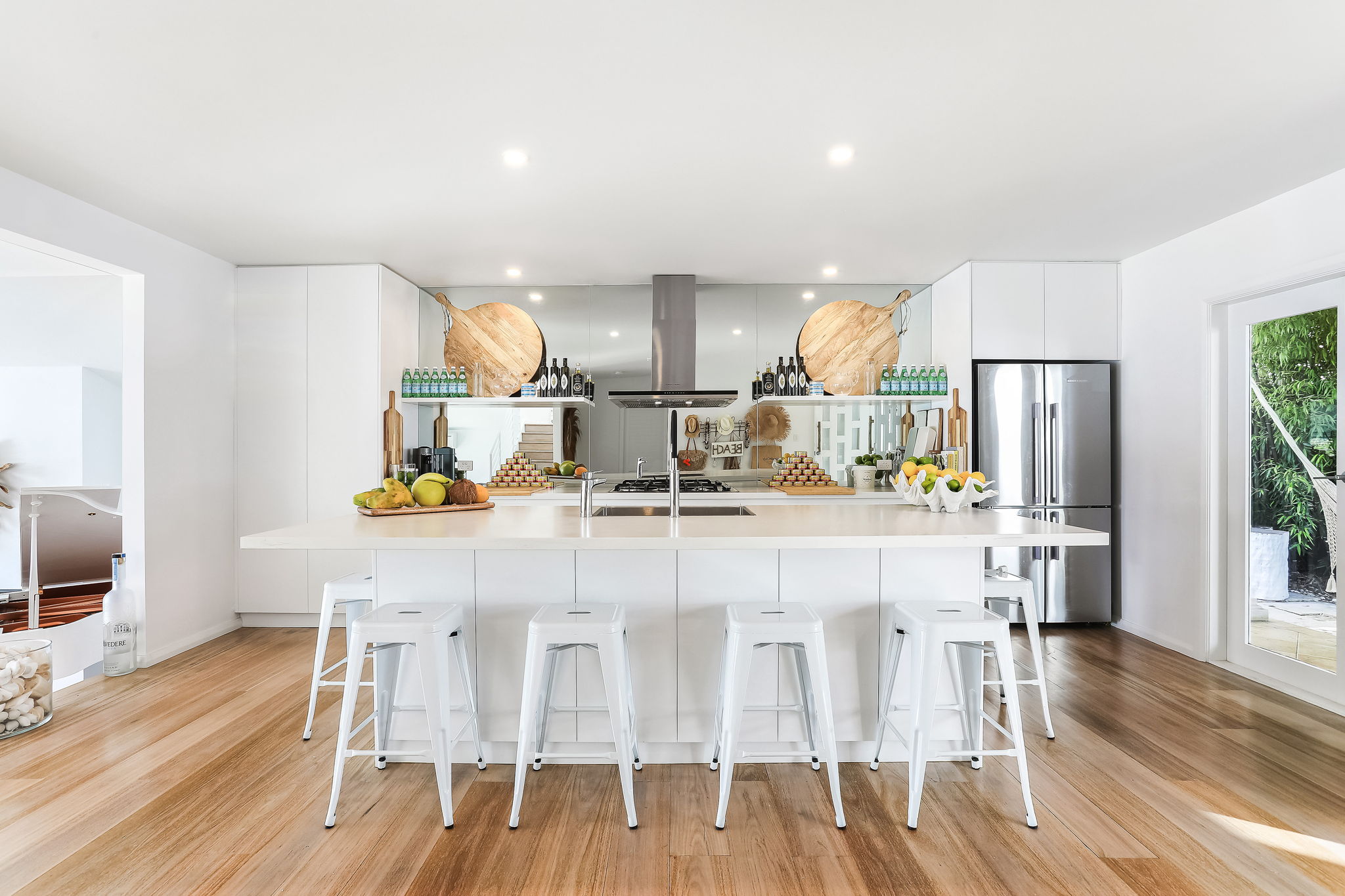 White kitchen island with four white stools, mirror splashback and timber floor