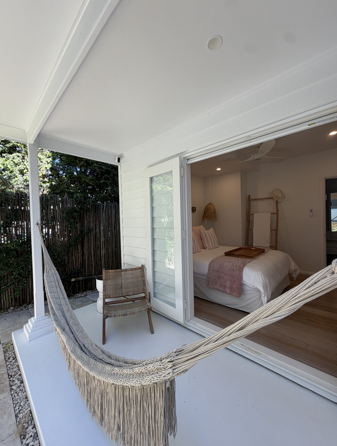 Fringed hammock on a white porch with wicker chair, bamboo fence and bedroom beyond