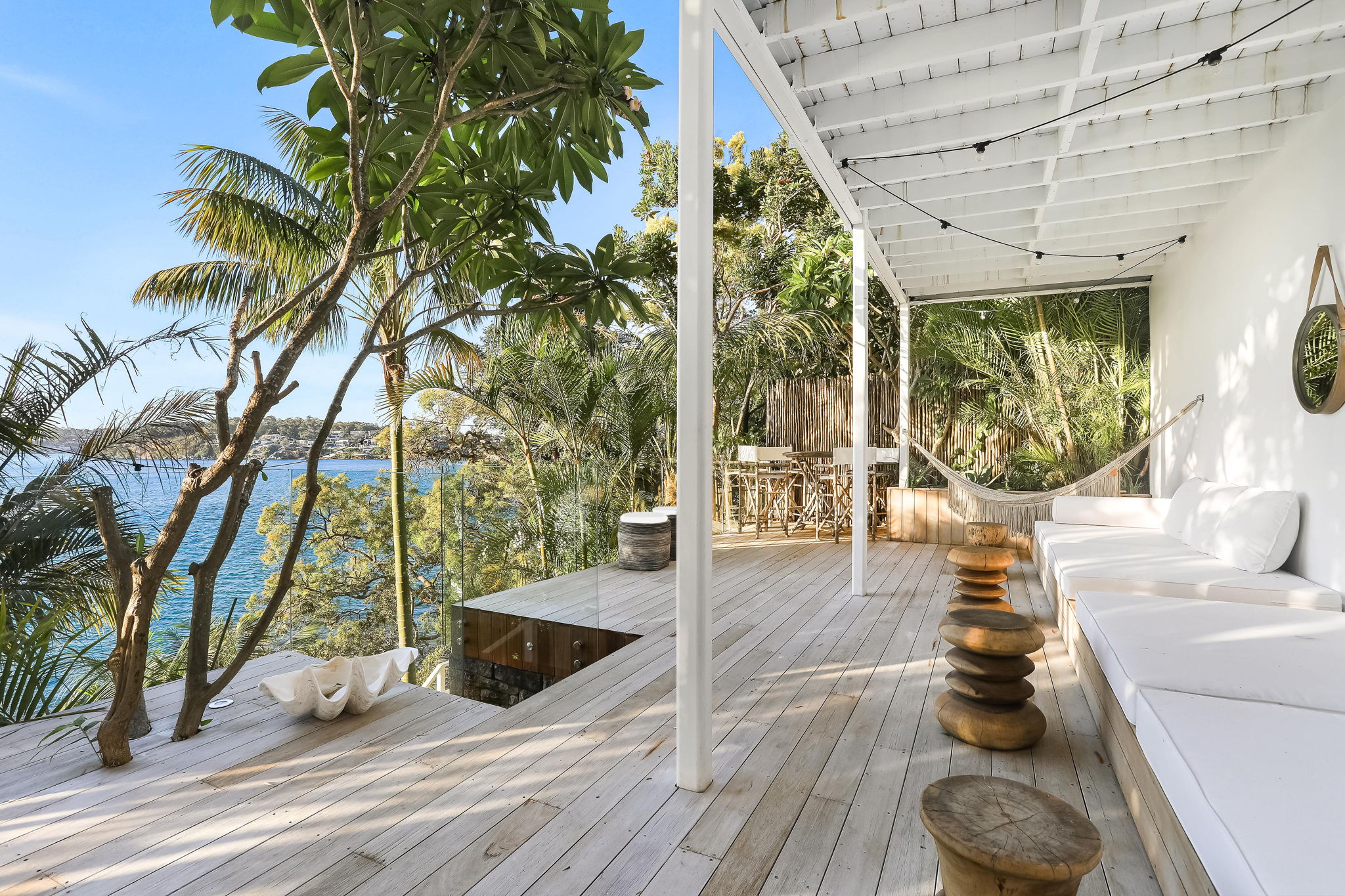 Waterfront deck with hammock, white daybed and stacked timber stools overlooking the bay
